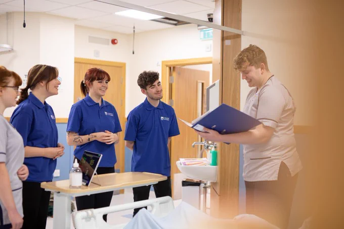 Photograph of student nurses in a clinical setting - five smiling students are gathered around a patient bed. One is reading out what appears to be clinical notes while the other four listen in