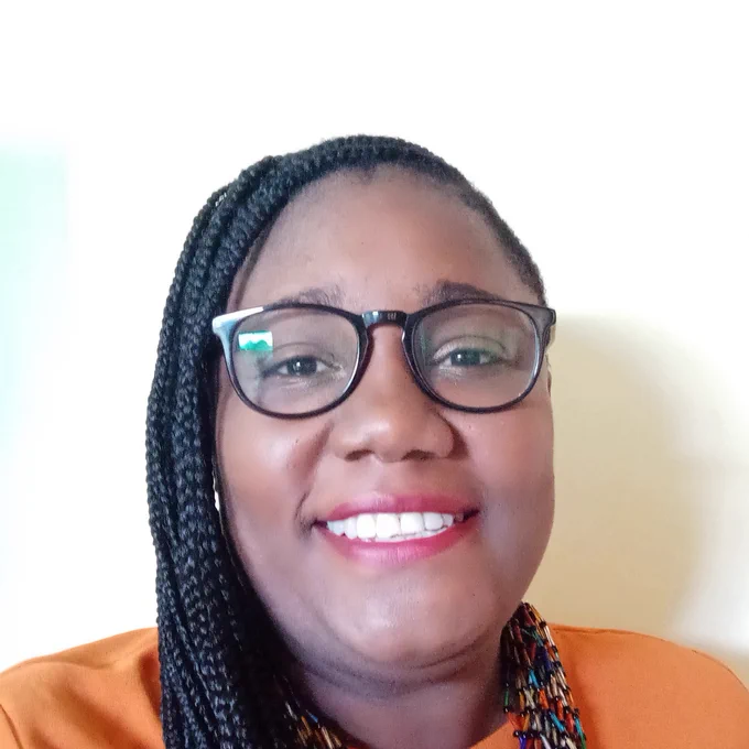 Amanda Thomas smiles at the camera. She has braided hair and is wearing an orange shirt and black glasses. 