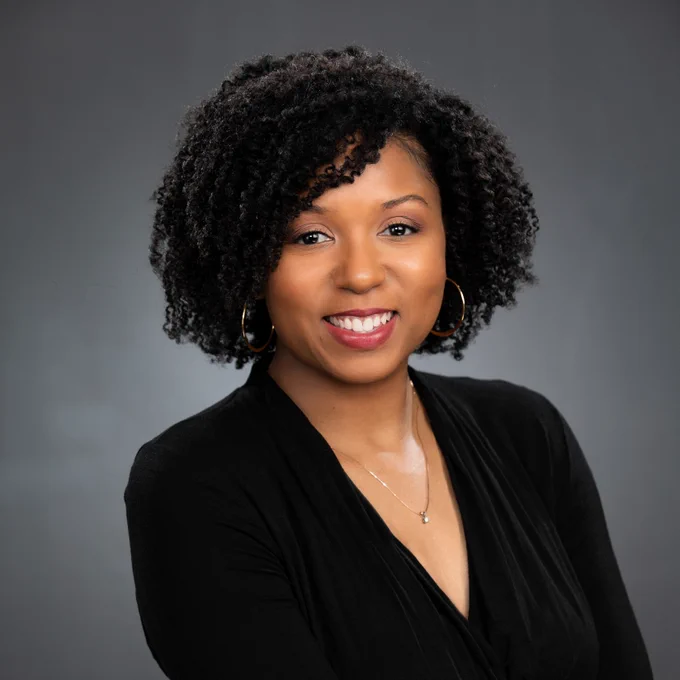 Lolita Tabron smiles at the camera in a studio with a grey background. She is wearin black clothes.