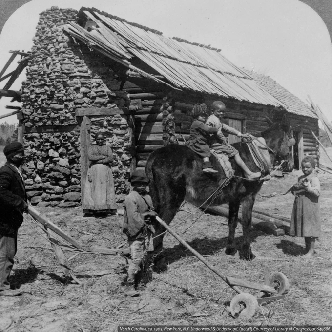A black and white photo of a stone and wood house with an African American family in front. Two children sit on a horse while the other two children plow. A man stands behind them with a plow, and a woman stands near the house door.