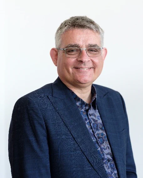 A photograph of Saul Nassé smiling in a navy suit and patterned shirt. 