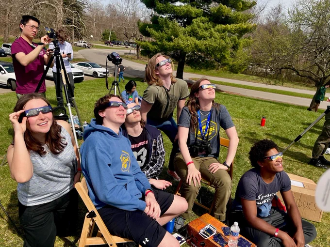 A group of six students watches the eclipse with their glasses on as another student sets up a camera.