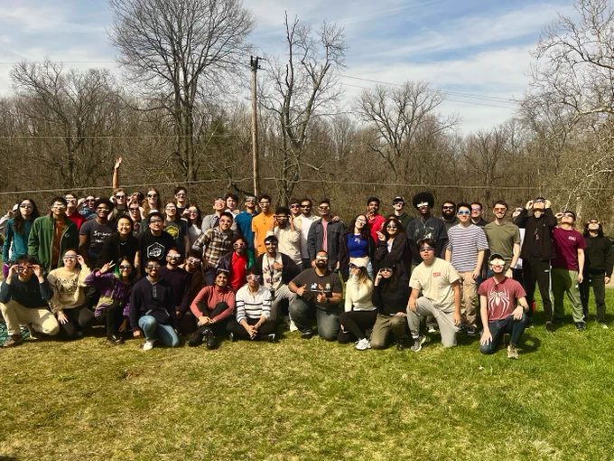 A large group of people stands on a grassy hill and smiles at the camera. Everyone is wearing eclipse glasses.