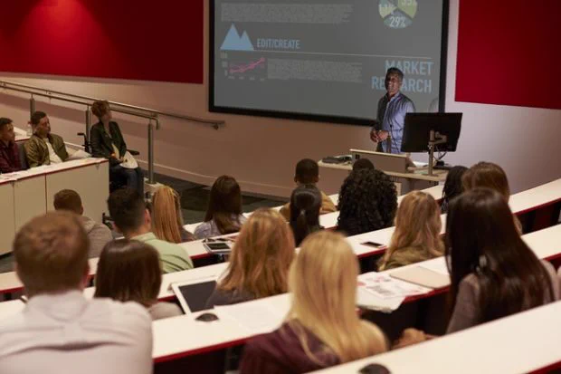 Students seated in a lecture hall watch as an academic lectures in front of large screen with graphs and graphics. 