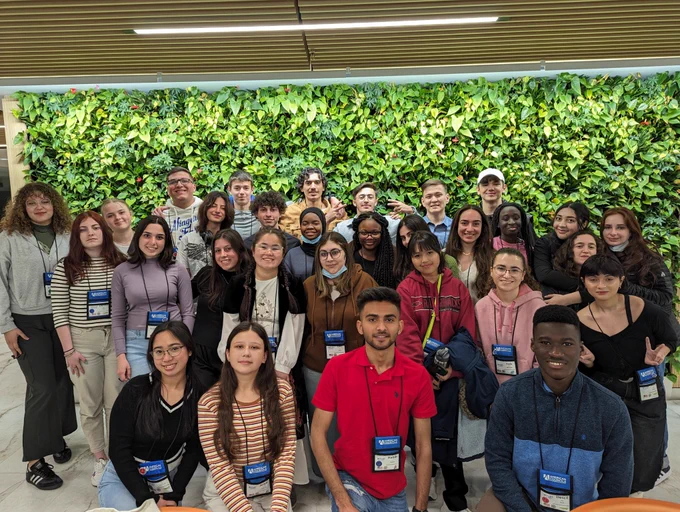 30 students pose as a group in front of a plant wall in an office