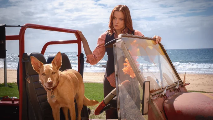 Anna Samson as MacKenzie Clarke standing by car with no roof or doors with a dog at the front looking to the camera. Beach and ocean in background