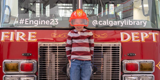 A photo of a child standing in front of a decommissioned fire truck. They are wearing a blue and red striped shirt, jeans and a fire fighter uniform helmet. 