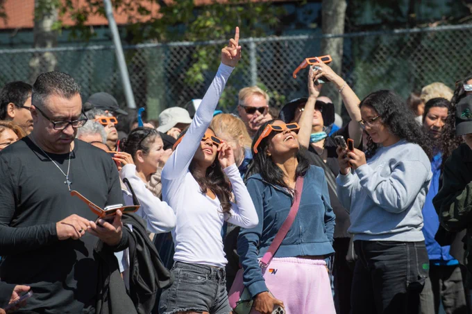 A crowd of people watches the eclipse at Caltech, a pair of friends smile and point up at the sky in the front of the group