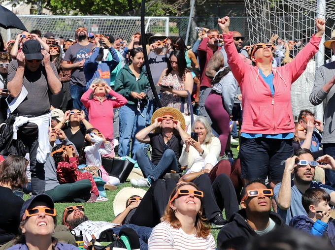 A crowd of people gather between the goals on a soccer field at Caltech's campus to watch the eclipse; at least one person raises their arms to cheer the event