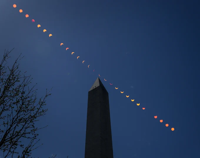 This composite image shows the progression of a partial solar eclipse over the Washington Monument in Washington. The orange Sun and lunar shadow track from the upper left to the lower right of the frame, across a dark blue sky and the pointed tip of the silhouetted obelisk. The bare branches of a tree reach into the lower left side of the frame. Credit: NASA/Bill Ingalls