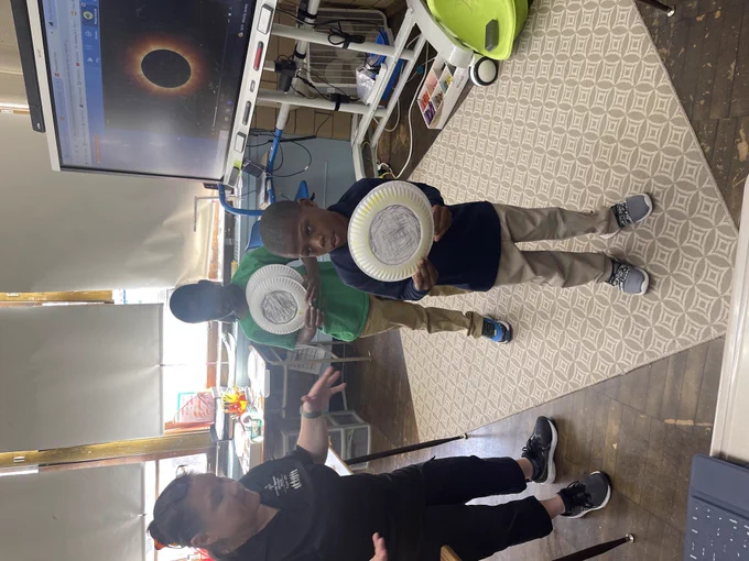 Two students stand next to their teacher presenting their paper plates they have colored to represent the 2024 solar eclipse 