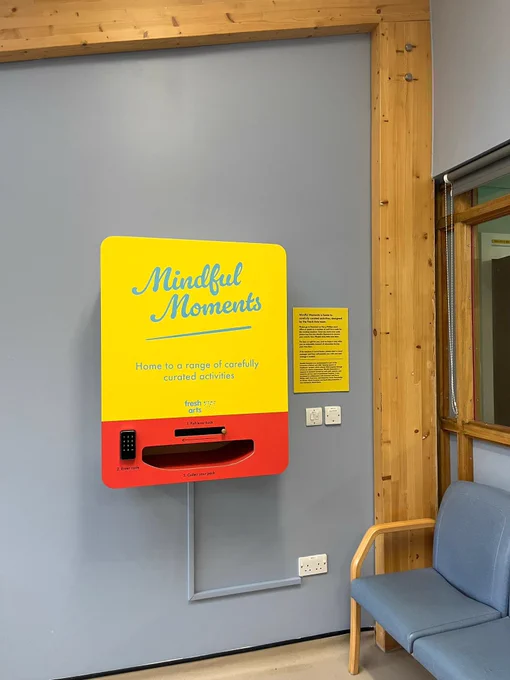 A photo of the mindful moments vending machine which is attacked to the walls with some seats next to it. It is bright yellow and red and has a handle to move and a gap to collect the activity packs from.