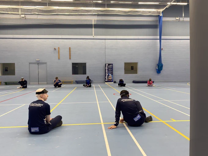The young people are sat on the floor of a sports hall. Two have their backs to the camera