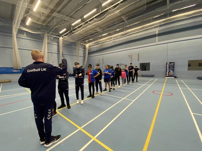 An activity is underway with all the participants stood in a line in a sports hall receiving instruction from Stephen