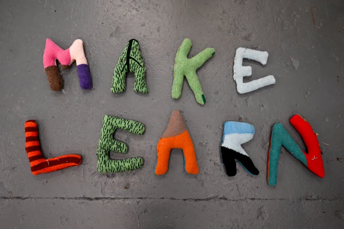 Colourful fabric letters spelling MAKE LEARN on a grey cement floor.