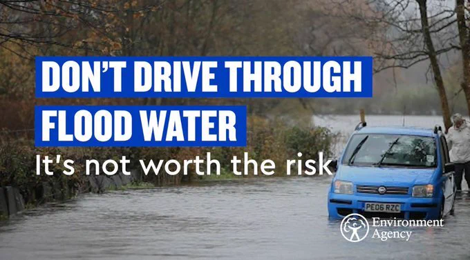 A blue car appears stuck on a flooded road. Water is up to the car's hub caps. The driver stands at the back of the vehicle. Water dominates the rest of the photo, with trees down the left-hand side.
