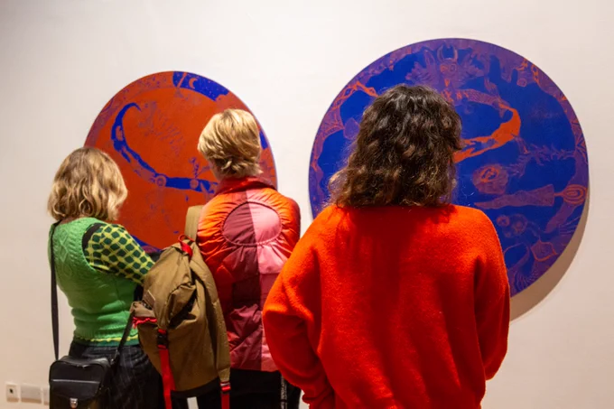 Three people stood in front of two circular paintings. One painting is orange with blue details and the other painting is blue with orange details. What the paintings are depicting can't be made out. The paintings are hung against a white wall.