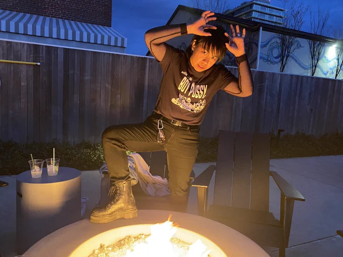 Sean standing in front of a fire pit, wearing all black. He wears a mesh long sleeve with a t-shirt over it that says, “boy pussy”, tucked into his jeans with a belt, and Doc Martens. Sean’s gaze is focused toward the camera as he gestures his hands above his head.
