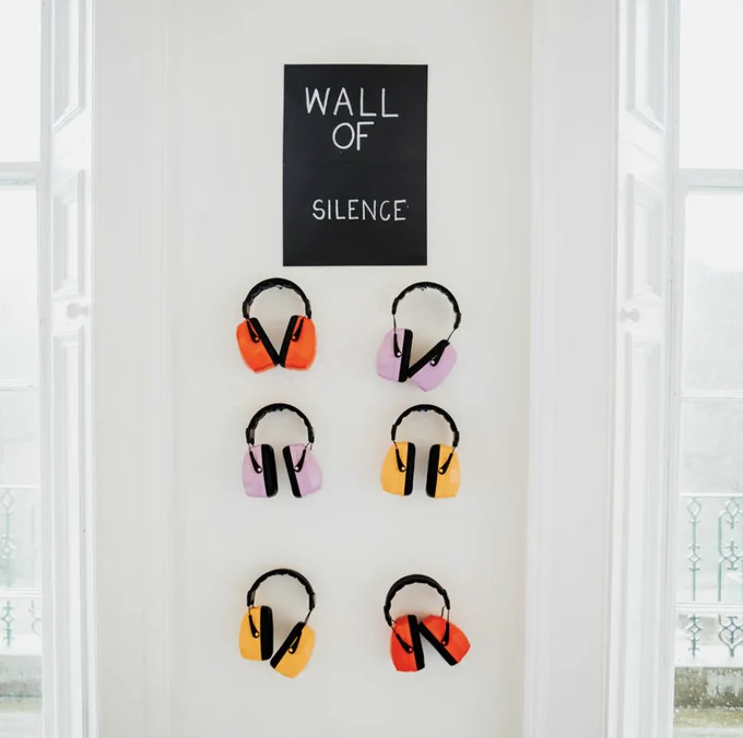 A white wall with a black sign that says 'Wall of Silence', above six pairs of brightly coloured headphones hanging underneath in red, lilac and yellow. 