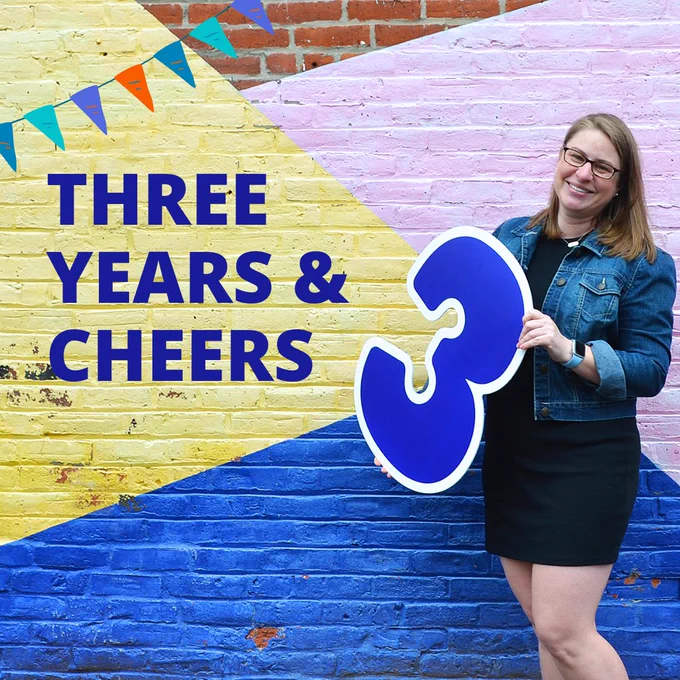 Elizabeth stands in front of a colorful geometric mural holding a large blue number three to celebrate their business anniversary.