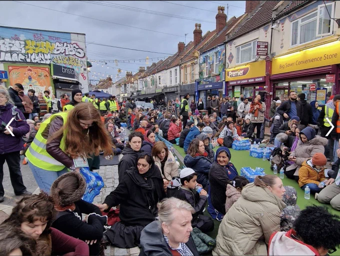 Many people sitting down together in the middle of St Mark's Road, waiting to be served food