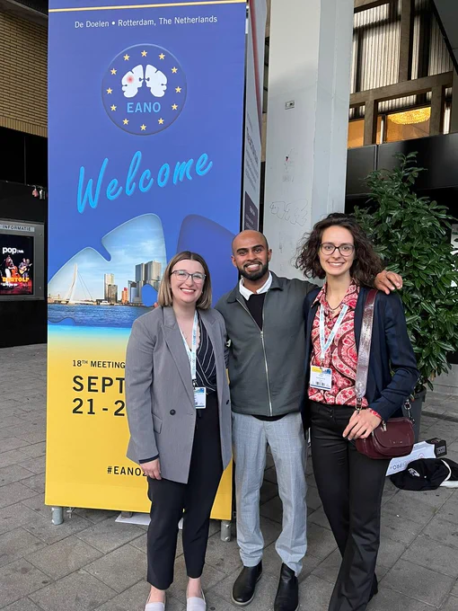 Graduate students Emma (left), Harshal (middle), and Helgi (right) standing in front of the European Association of Neuro-oncology conference banner in Rotterdam, Netherlands.