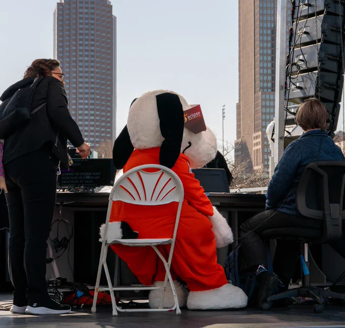 Snoopy, who is wearing an orange NASA flight suit and red NASA eclipse glasses, sits in a chair at the NASA broadcast desk. The Cleveland skyline can be seen in the distance. Image Credit: NASA/Quentin Scwhinn
