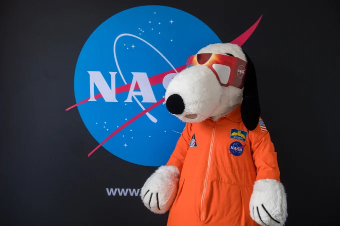 Snoopy, who is wearing an orange NASA flight suit and red NASA eclipse glasses, poses in front of a large sign featuring the NASA "meatball" insignia. Image Credit: NASA/Jef Janis