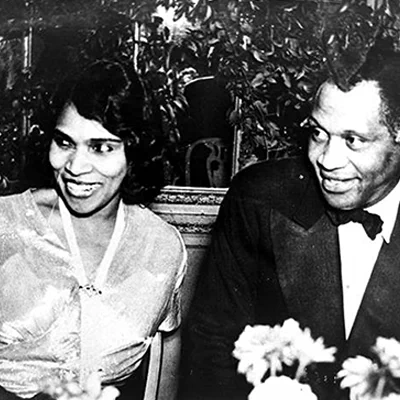 1946 photo of Marian Anderson on the left sitting with Paul Robeson in a tuxedo looking and smiling at something out of shot. This was a dinner party held in her honor. 