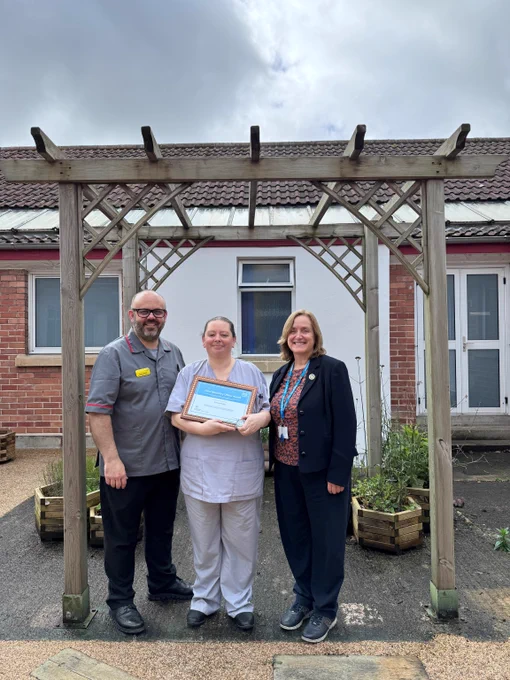 A person in grey scrubs holding a certificate with two others either side of her.-