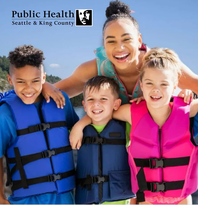 Four kids are smiling, embracing, and posing for the camera at the beach on a sunny day. They are wearing life jackets. The King County logo is in the upper left.