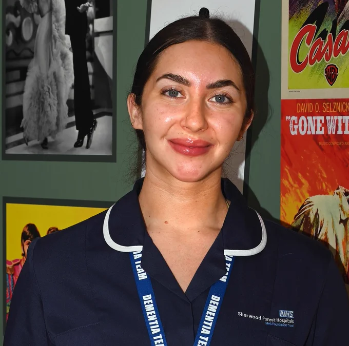A photograph of a nurse in uniform standing in front of posters for old movies such as Casablanca and Gone With the Wind. 