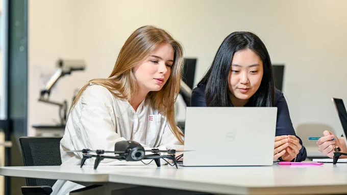 Two female computer science students work together on a laptop with a drone beside it