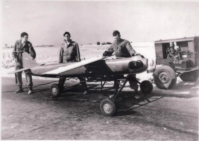 Three men stand in an old black and white photo standing around an operational a remote control aerial target.