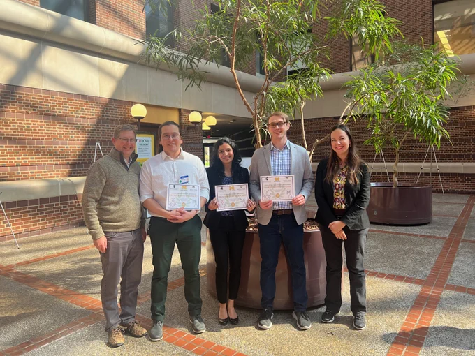 MSU chemistry graduate students pose with their awards alongside  department faculty. The photo is taken in a brick and stone courtyard with two potted trees and brick building in the background.