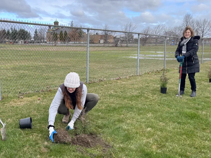 Volunteer planting tree and another volunteer holds a shovel next to a small tree in a pot.