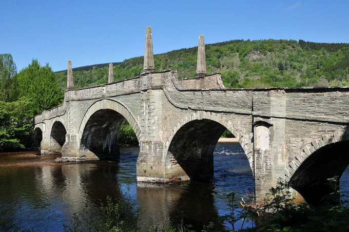 The Tay Bridge at Aberfeldy. The image shows an ornate five arched stone bridge obliquely crossing a river with the right-hand end starting from the near bank. The central arch is decorated by tapering stone columns set into the parapet. Beyond the river is a tree-covered hillside.