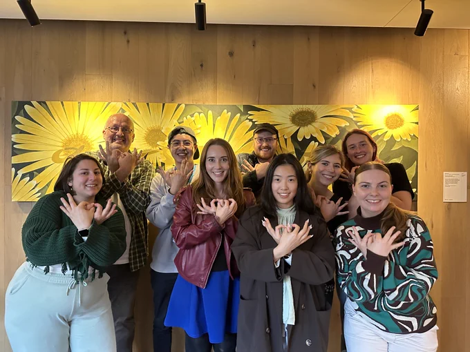 An image of nine smiling people in front of a poster of sunflowers. They are making bee symbols with their hands.