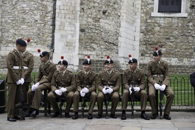 A group of soldiers sit on a bench. They are all dressed in their brown uniforms and have red and white hackles on their berets.