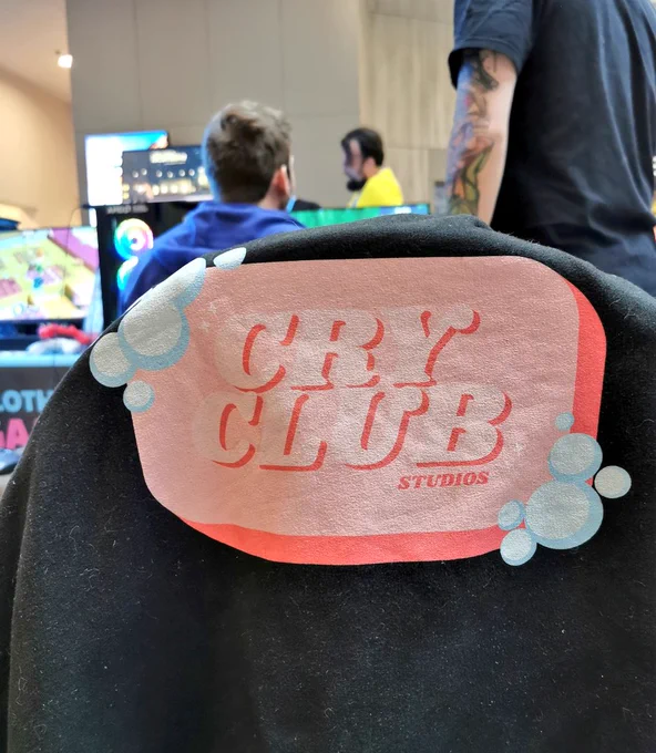 A photo of a hoodie on a chair, the logo is soap shaped and says Cry Club Studios. There are people behind it playing games and talking. 