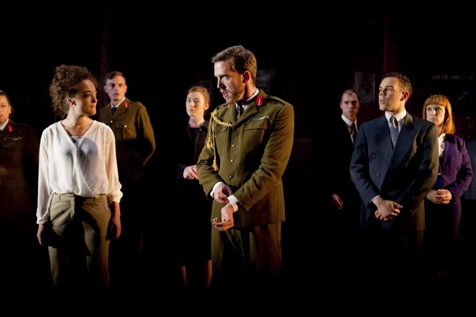 A group of people stand spaced out on stage. A man wearing a army generals uniform stands in front sorting out his shirt cuff. Some people around him also wear army general uniform and others wear suits.
