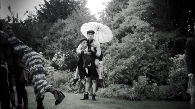A man gives another man a piggy bag ride. The man on top is holding a white umbrella. They are outdoors in a garden. The colours in the image are faded. They are wearing shirts jumpers and shorts.