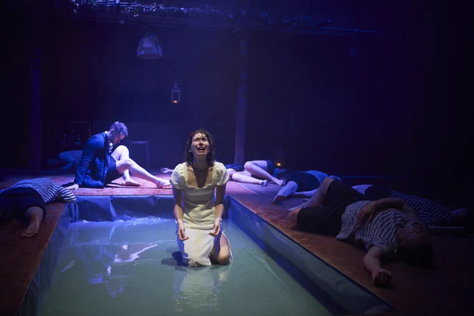 A girl is on her knees in a shallow pool crying. A man ponders on the floor a feet behind her. Various people wearing black and white stripped T-shirts lay dead around them.
