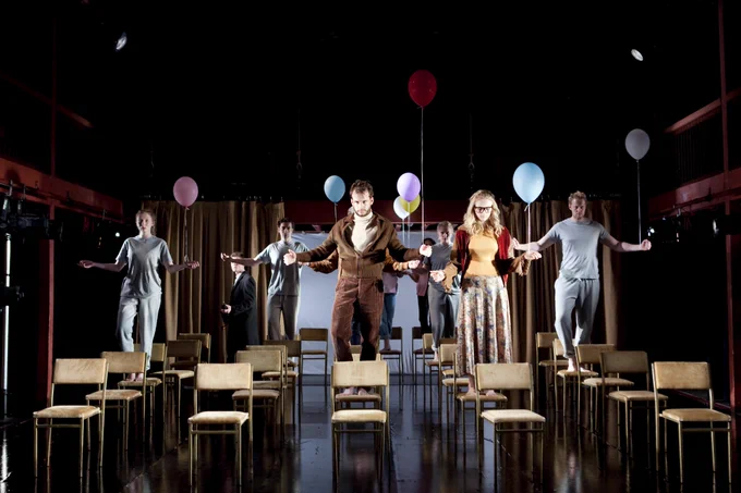 Five rows of light brown chairs are on a stage. A group of actors are standing on some of the chairs holding a balloon in their left hand. They have their arms spread and all look ahead. Some are wearing light blue pyjamas. The man in front wears a brown jacket and brown corduroy trousers.