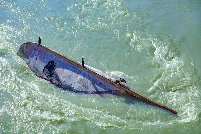 Photo of a capsized boat surrounded by shallow green-blue ocean water. The sharp line of the boat's overturned bottom resembles the spine of an animal, and there are three black seabirds perched on it.