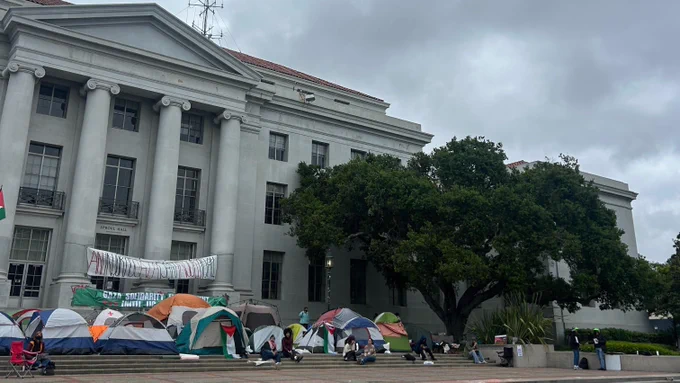 A photo of the Free Palestine Encampment on Sproul Plaza. Several multicolored tents are in front of Sproul Hall. Two banners behind the tents read "An injury to Gaza is an injury to all" and "Gaza solidarity encampment until UC divests."