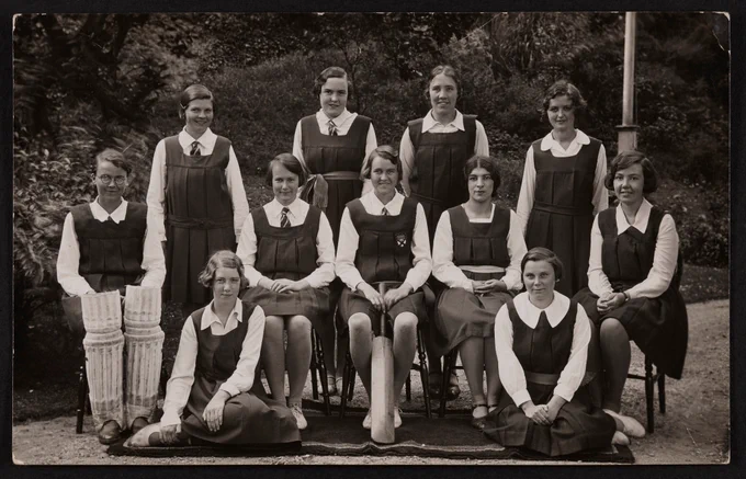 Black and white formal photograph of 11 women: four standing, five seated on chairs, and two seated on the ground. They are wearing white blouses and dark pinafore dresses. The woman seated in the centre is holding a cricket bat and the woman seated on the left is holding two knee pads.