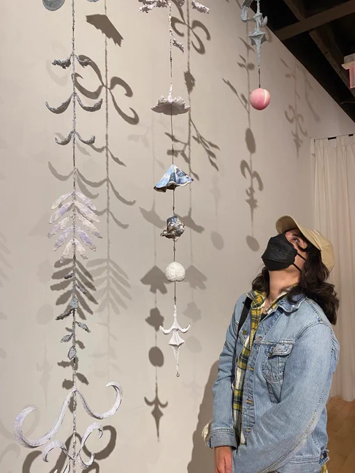 [Image Description: a photo of a visitor to Hamilton Artists Inc viewing installation by Sangmin Lee. The visitor is wearing a blue denim jacket and a black KN95 mask. The installation is a collection of hanging figures made of plaster and paper and foam hanging from the ceiling of the gallery.]