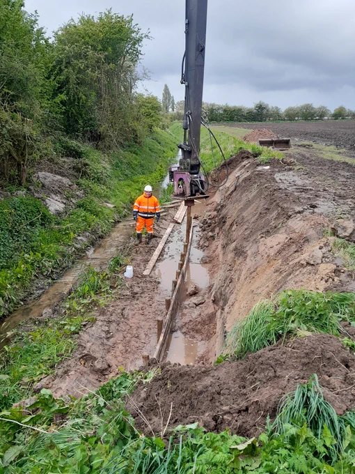 Operative stood in watercourse while machine operates to repair bank slip