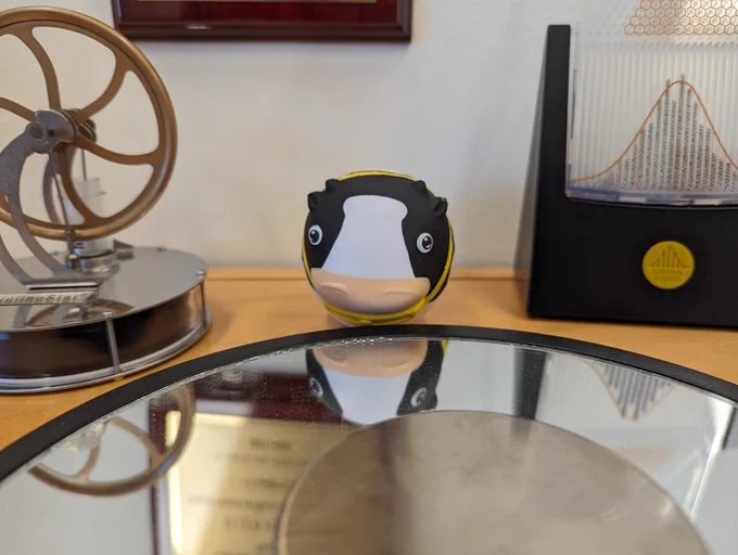 Photo of a spherical cow squeeze ball sitting next to a Galton board, a Stirling engine, and an Euler disk.